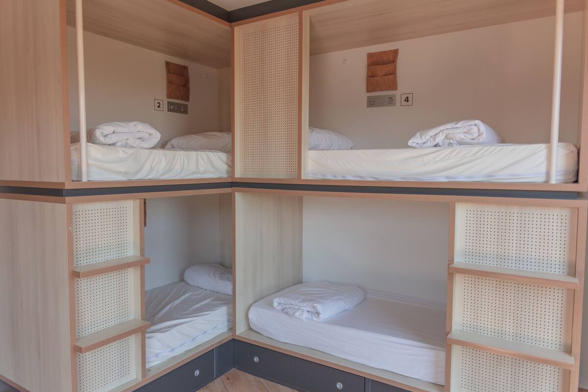 A four-bedded room with wooden frames and a central divider. Each bed has a white mattress and pillows, and there are storage drawers under the beds.