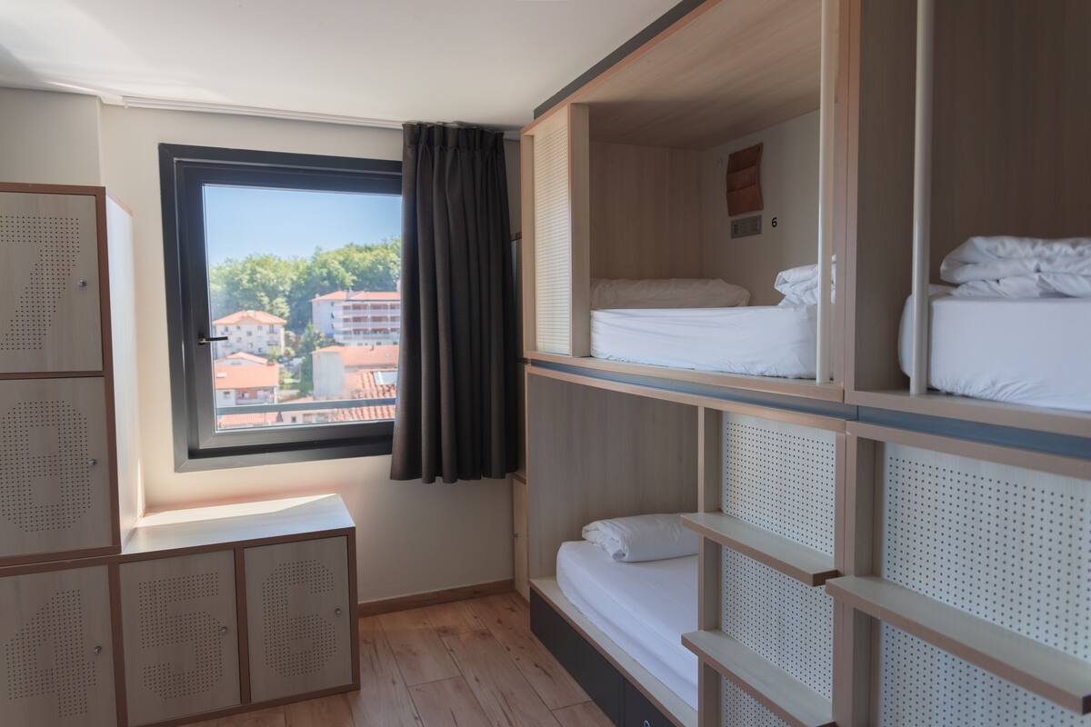 A room with bunk beds, a window with a view of buildings, and a wooden cabinet.