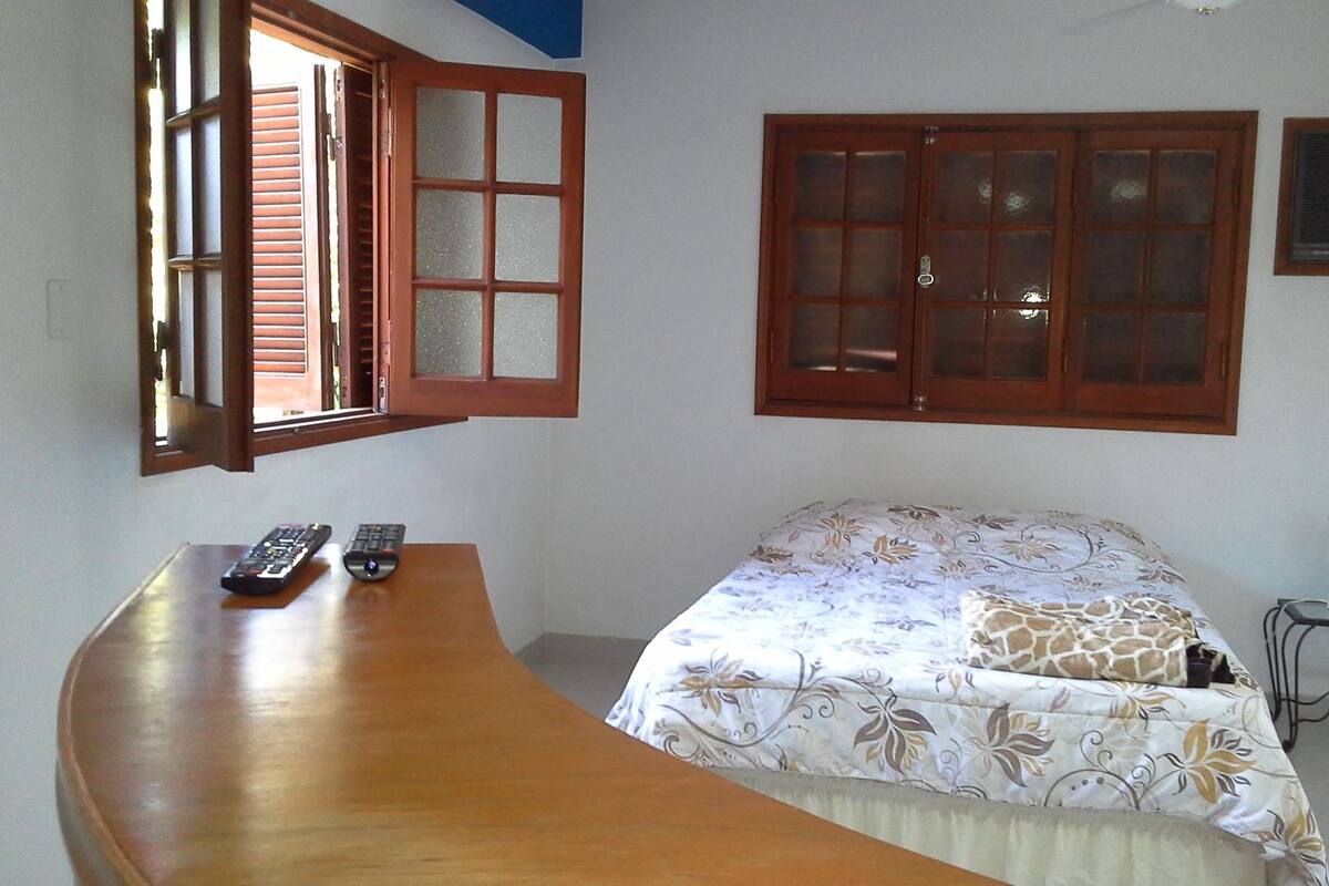 A bedroom with a bed, a desk with two remote controls, a window with open shutters, and a closed door.
