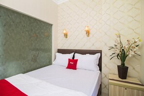 A neatly made bed with white bedding and a red pillow, a wall with a geometric pattern, and a potted orchid plant on a bedside table.