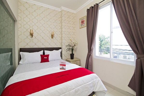 A neatly made bed with a red accent pillow, a small bedside table with a plant, and a window with curtains.