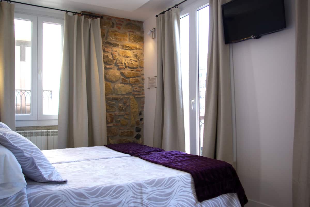 A bedroom with a stone wall, a bed with white bedding and a purple blanket, a TV mounted on the wall, and a window with curtains.