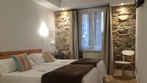 A bedroom with two beds, a stone wall, a window with curtains, a small table with a lamp, and a chair. - Pension Balerdi (San Sebastián)
