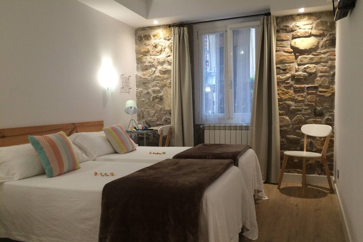 A bedroom with two beds, a stone wall, a window with curtains, a small table with a lamp, and a chair.