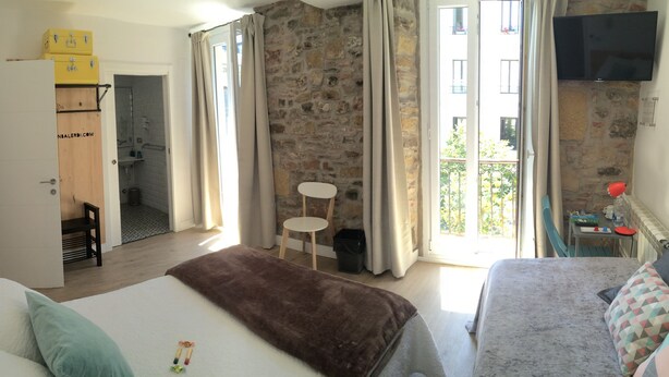 A hotel room with a stone wall, a bed with a brown blanket, a sofa with patterned cushions, a small table, and a TV mounted on the wall. - Pension Balerdi (San Sebastián)