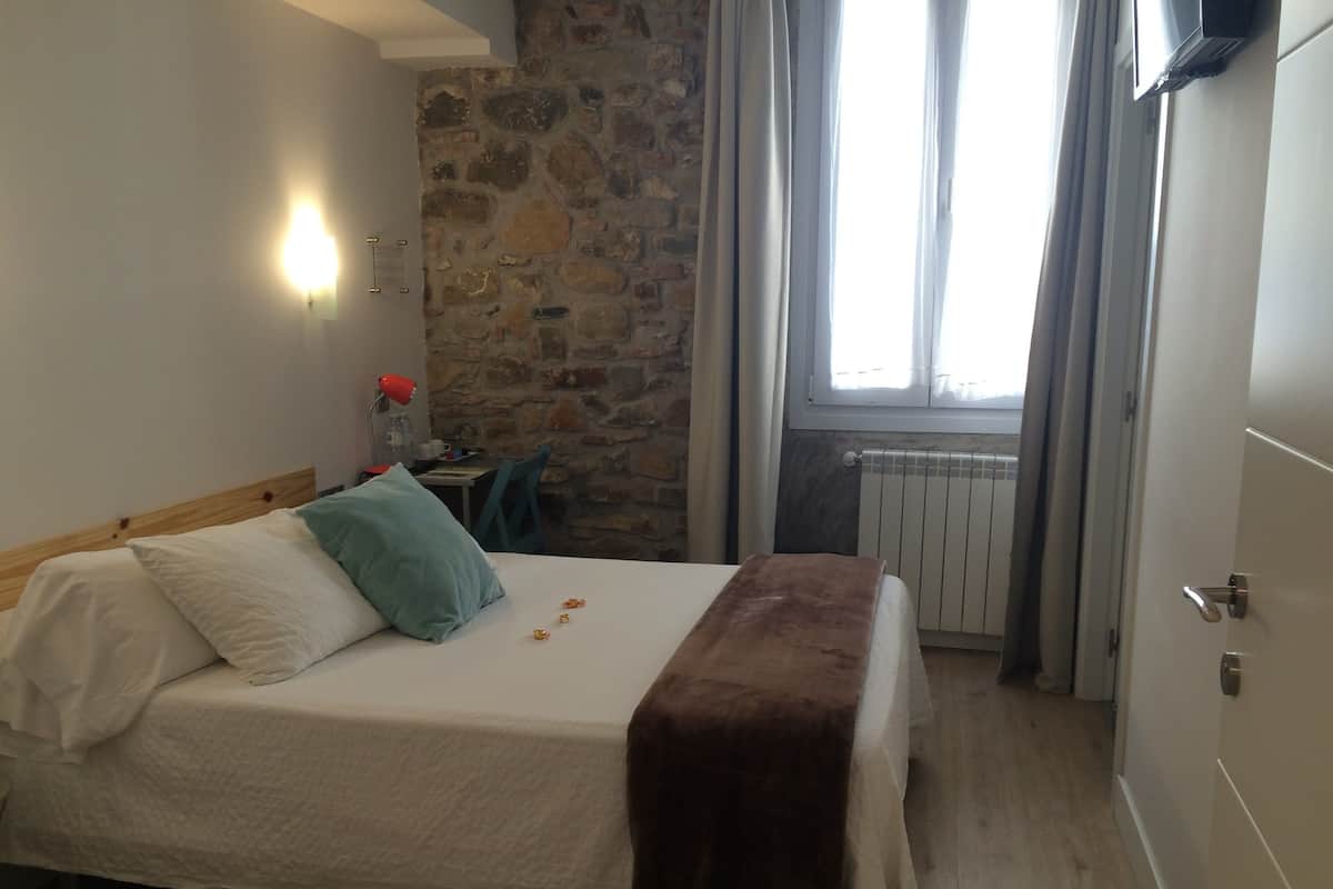 A bedroom with a stone wall, a bed with a white bedspread, a brown throw blanket, and a window with curtains.
