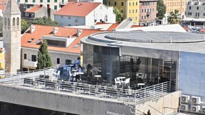 Een terras op het dak met buitenmeubilair, een modern gebouw met een gebogen glazen gevel, en op de achtergrond traditionele gebouwen met rode pannendaken.
