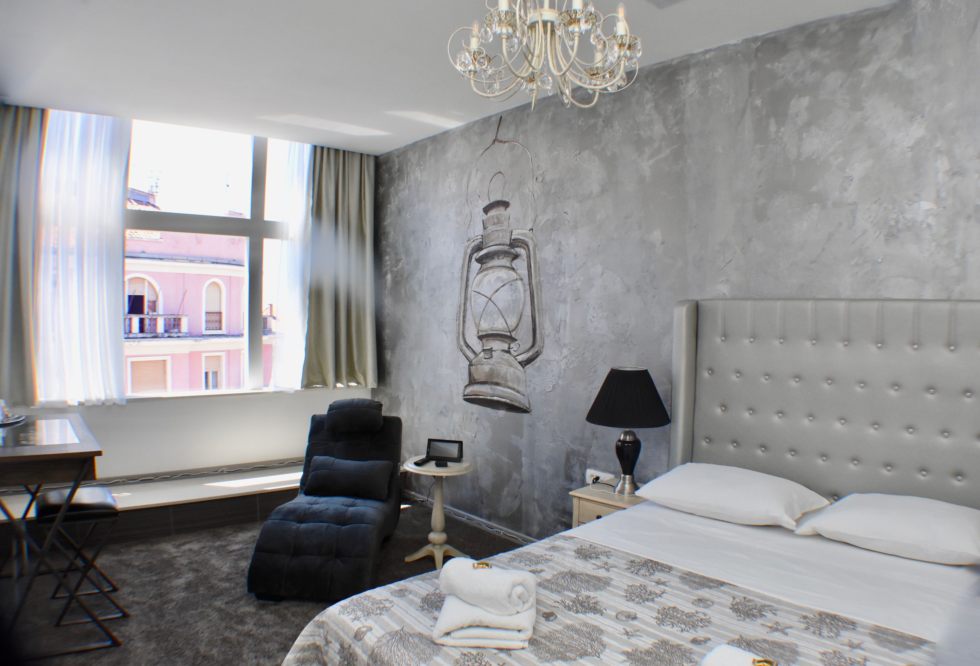 Quarto de hotel com uma cama grande, uma cadeira, uma mesinha, um abajur e vista para um edifício pela janela.