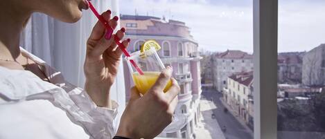 A person holding a drink with a lemon slice and a straw, looking out from a window with a view of buildings.