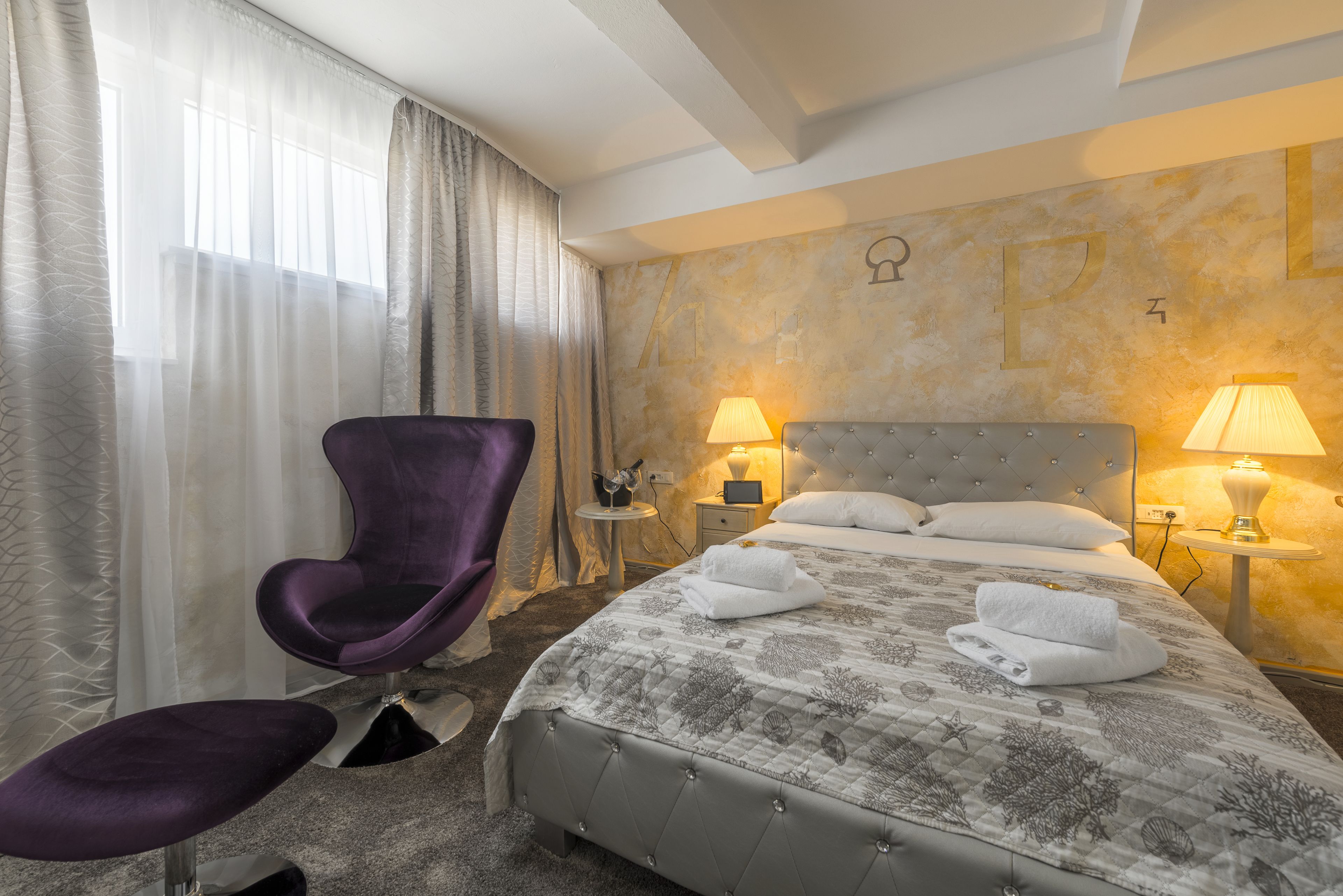 Habitación de hotel con cama, una silla morada, dos mesitas de noche con lámparas y una ventana con cortinas de gasa.
