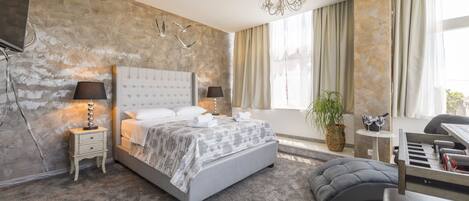 Un dormitorio moderno con una cama grande, una mesita de noche con lámparas, una pequeña mesa auxiliar, una planta en maceta y una zona de descanso con una silla y una mesa.