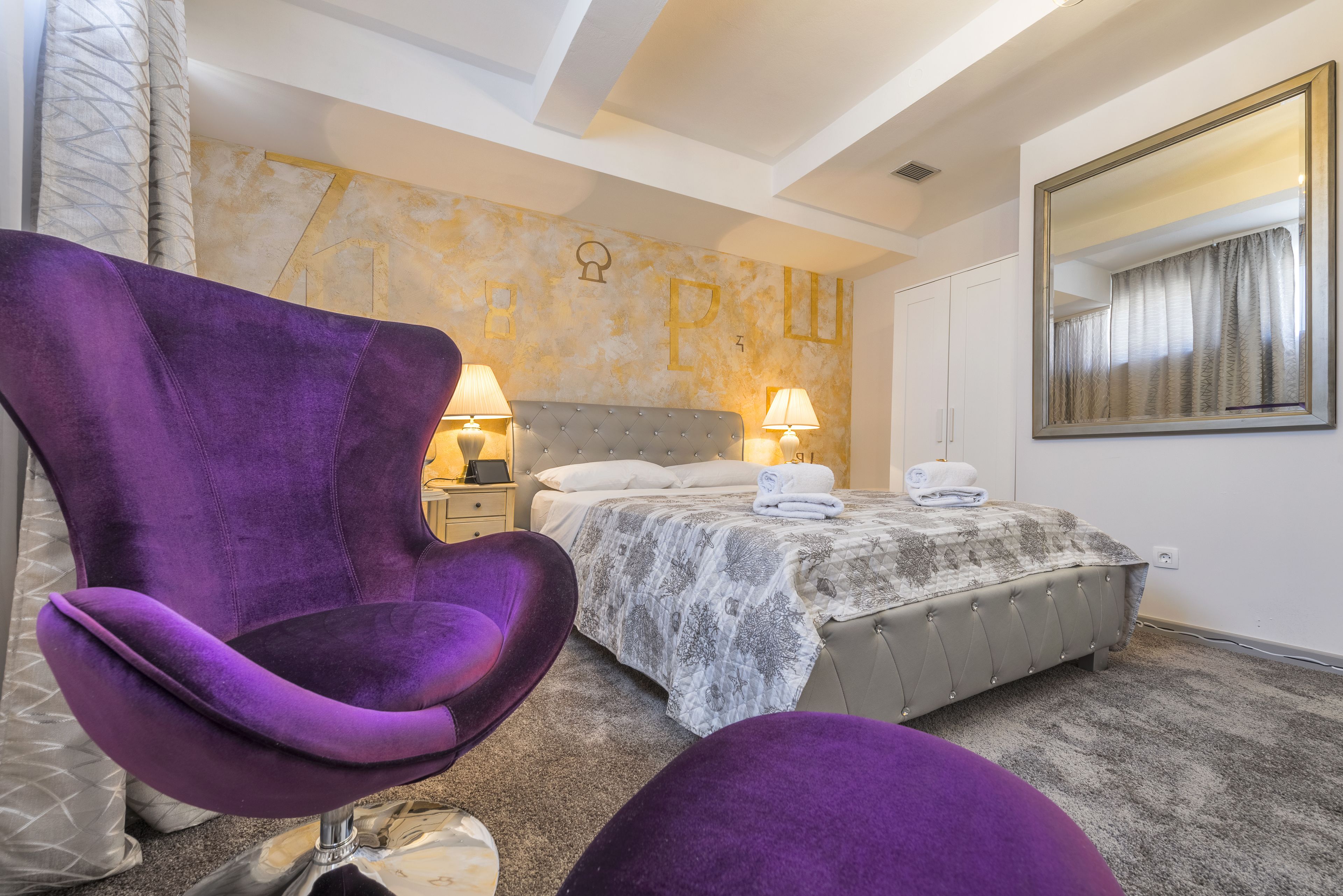 A bedroom with a bed, two nightstands, a purple armchair, and a large mirror.