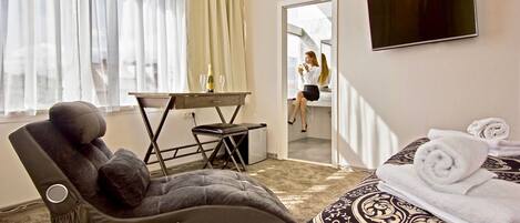 A hotel room with a massage chair, a bed with rolled towels, a TV on the wall, and a window with curtains.