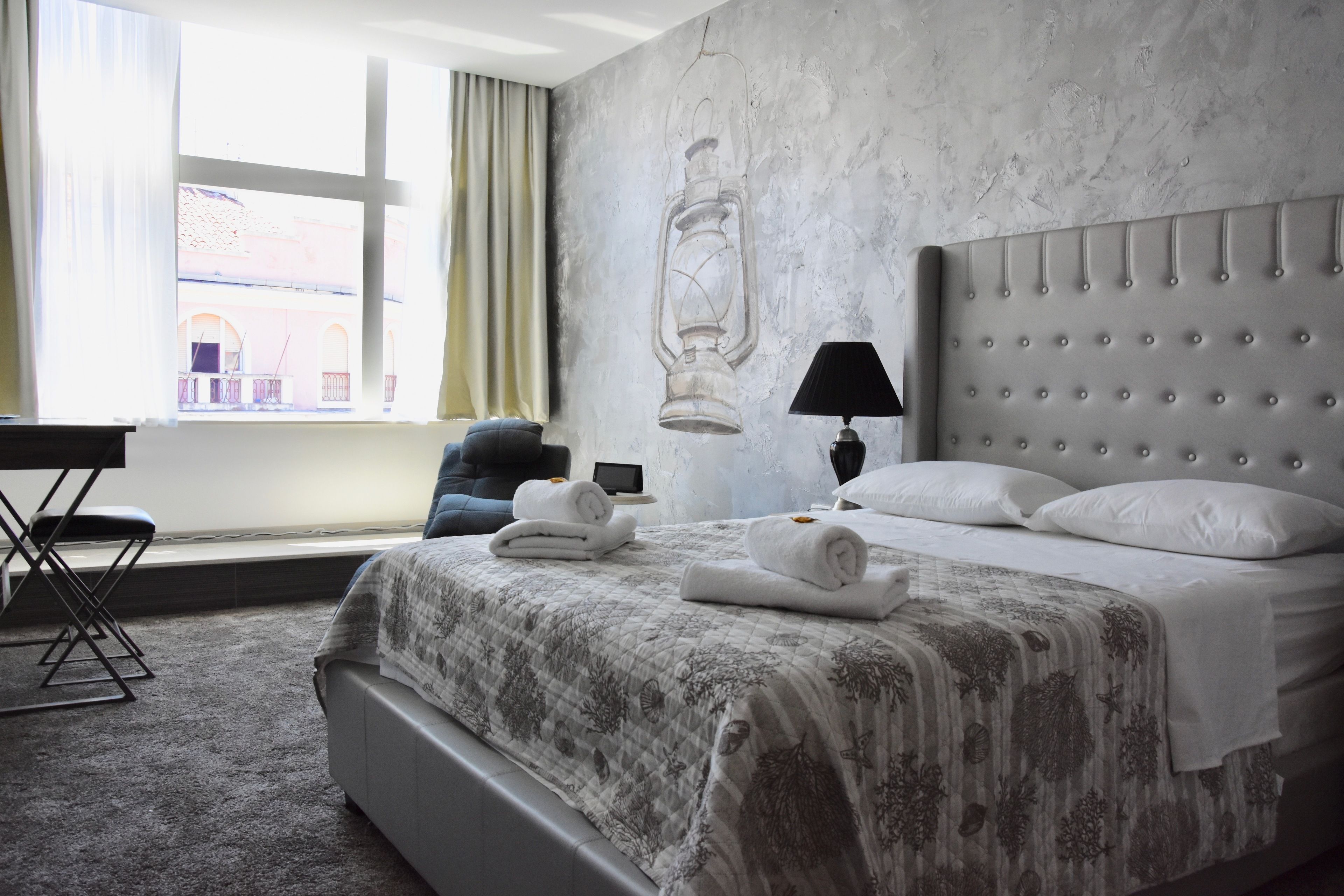A hotel room with a bed, a desk, a chair, a lamp, and a view of the outside.
