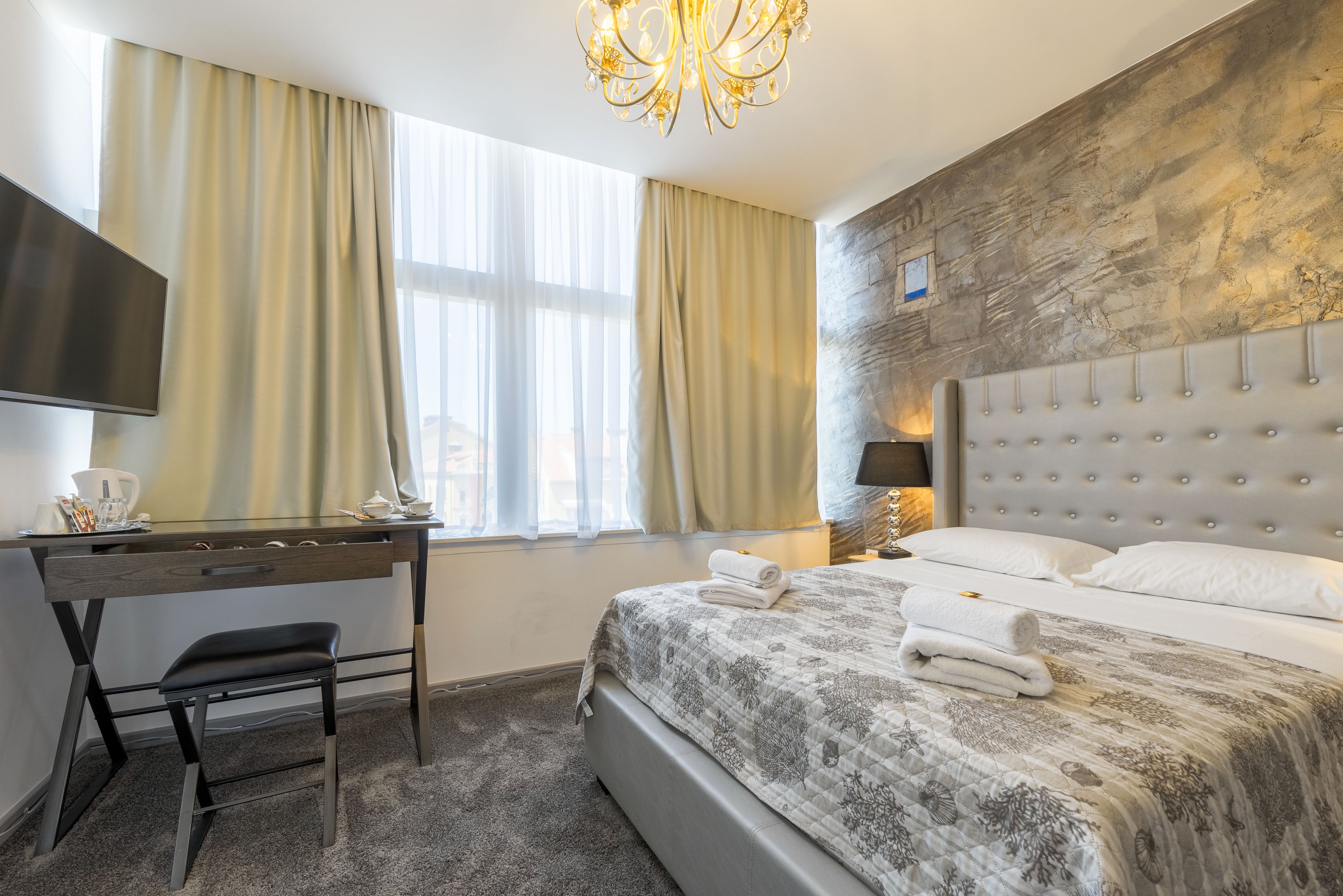 A hotel room with a bed, a desk, a chair, a television, and a chandelier.