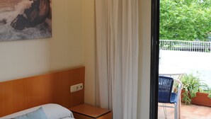 A hotel room with a bed, a nightstand, a painting of a beach scene, a view of a balcony with plants and a chair, and a sliding glass door. - Hotel Aitana (Irun)