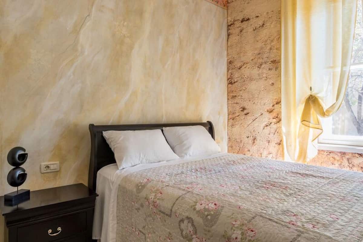 A bedroom with a bed, a nightstand, a wall socket, and a window with curtains.