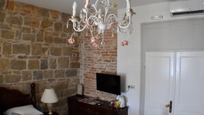 Una habitación con pared de piedra, una puerta blanca, un televisor sobre un mueble bajo, un aparador de madera y una araña decorada con rosas.