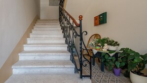 Una escalera con peldaños blancos y barandillas de hierro negro, que conduce a un rellano con una mesa y plantas en macetas.