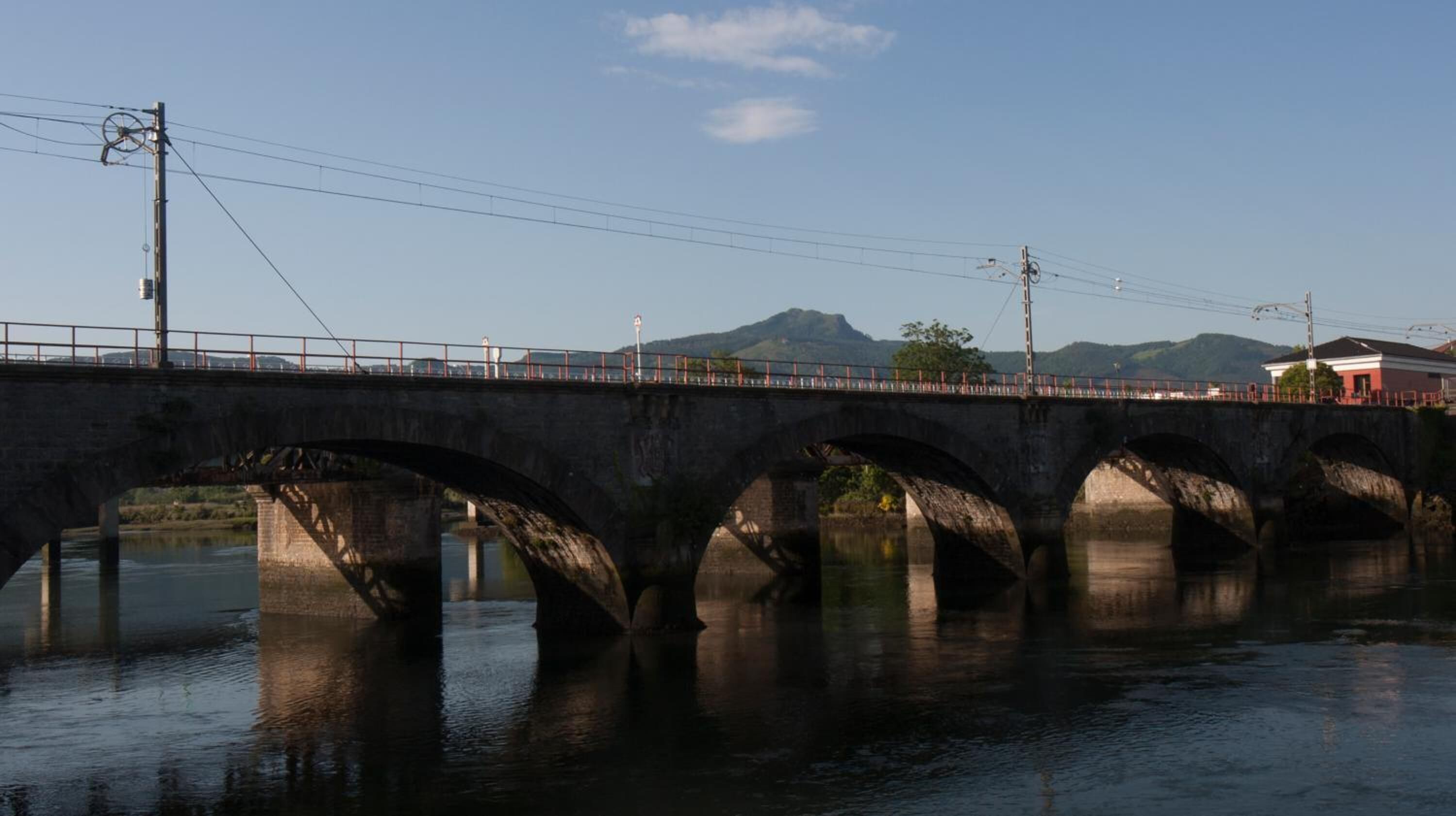 Un puente de piedra con múltiples arcos que cruza un río tranquilo, con un cielo despejado y colinas lejanas al fondo.