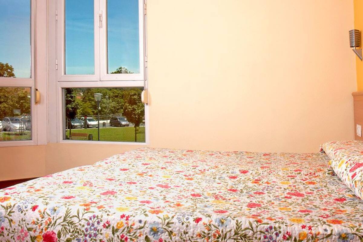 Un dormitorio con colcha estampada, ventana con vistas a coches aparcados y zonas verdes, y una lámpara fijada a la pared.