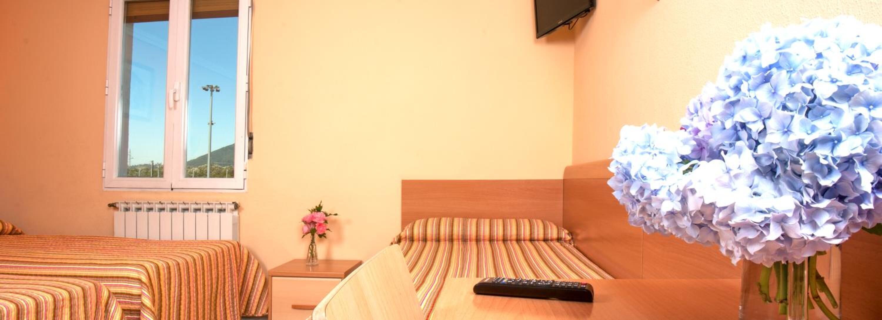 A hotel room with two beds, a nightstand, a vase with flowers, and a window with a view of the outside.