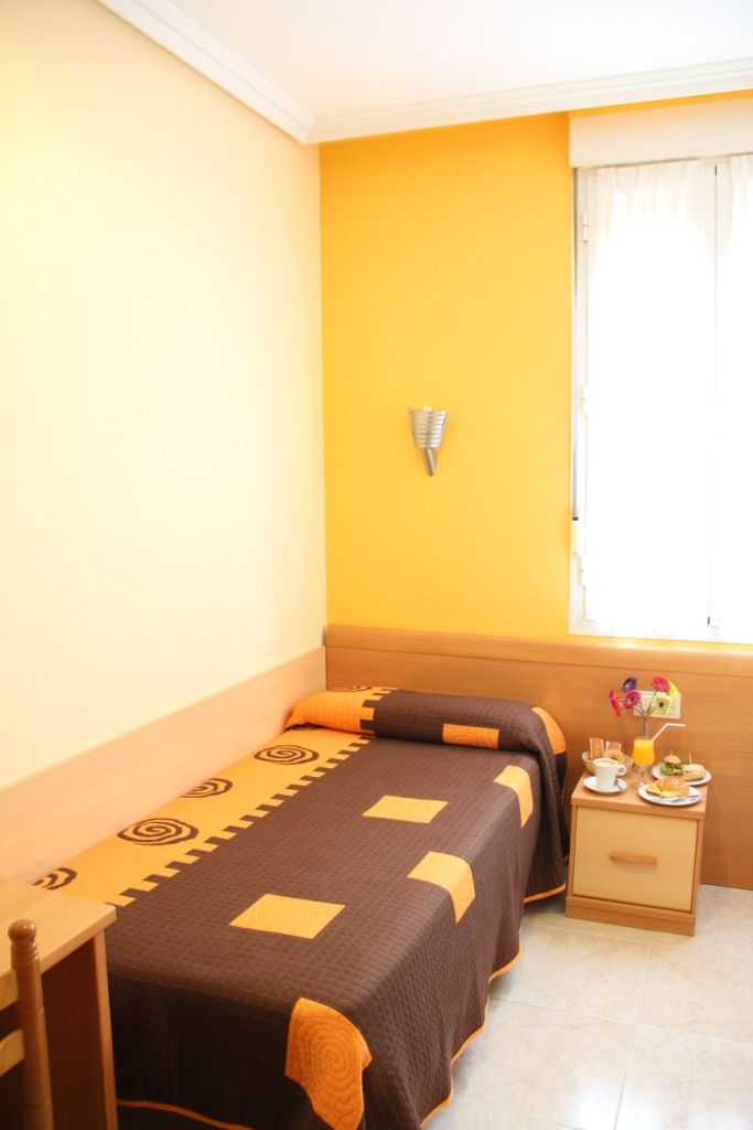 Una habitación pequeña con cama individual, pared amarilla, ventana con cortinas blancas, mesita de noche con una taza y un plato de comida, y una lámpara de pared.