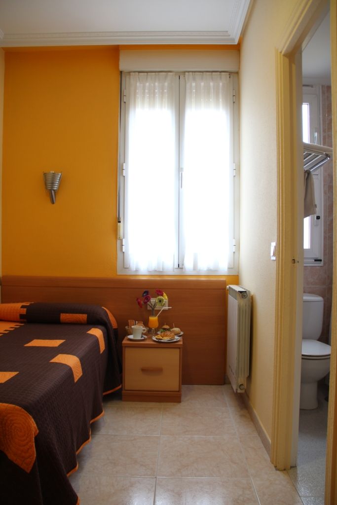 A hotel room with a bed, a bedside table with a vase of flowers, a radiator, and a window with curtains.