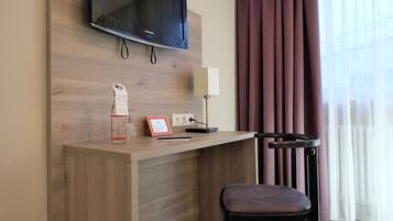 A hotel room with a wooden desk, a television mounted above it, a lamp, a chair, and a window with curtains.