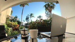 A laptop on a glass table with a white mug and a silver pot, reflecting palm trees and a clear sky.