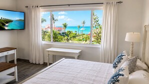 A bedroom with a large bed, a desk, a TV, and a window with a view of a beach and ocean.