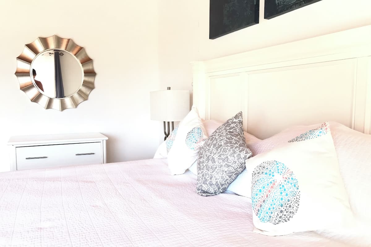 Een netjes opgemaakt slaapkamer met een bed, kussens, een nachtkastje, een lamp en een spiegel aan de muur.