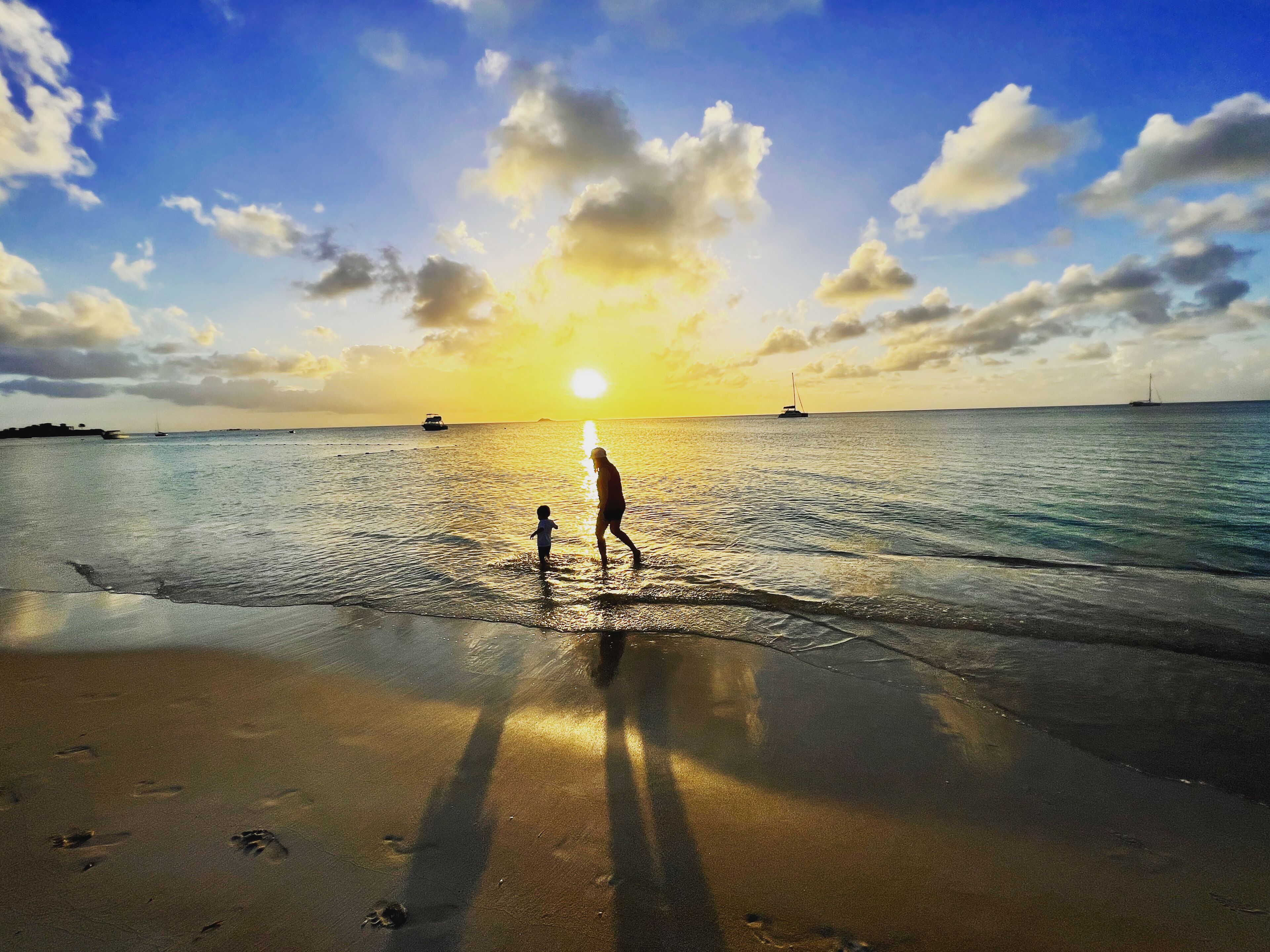 Escena de playa con un atardecer, mar tranquilo y una persona caminando con un niño.