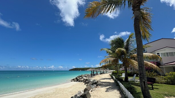 A beach with a clear sky, a sandy path, palm trees, and a building with a balcony.