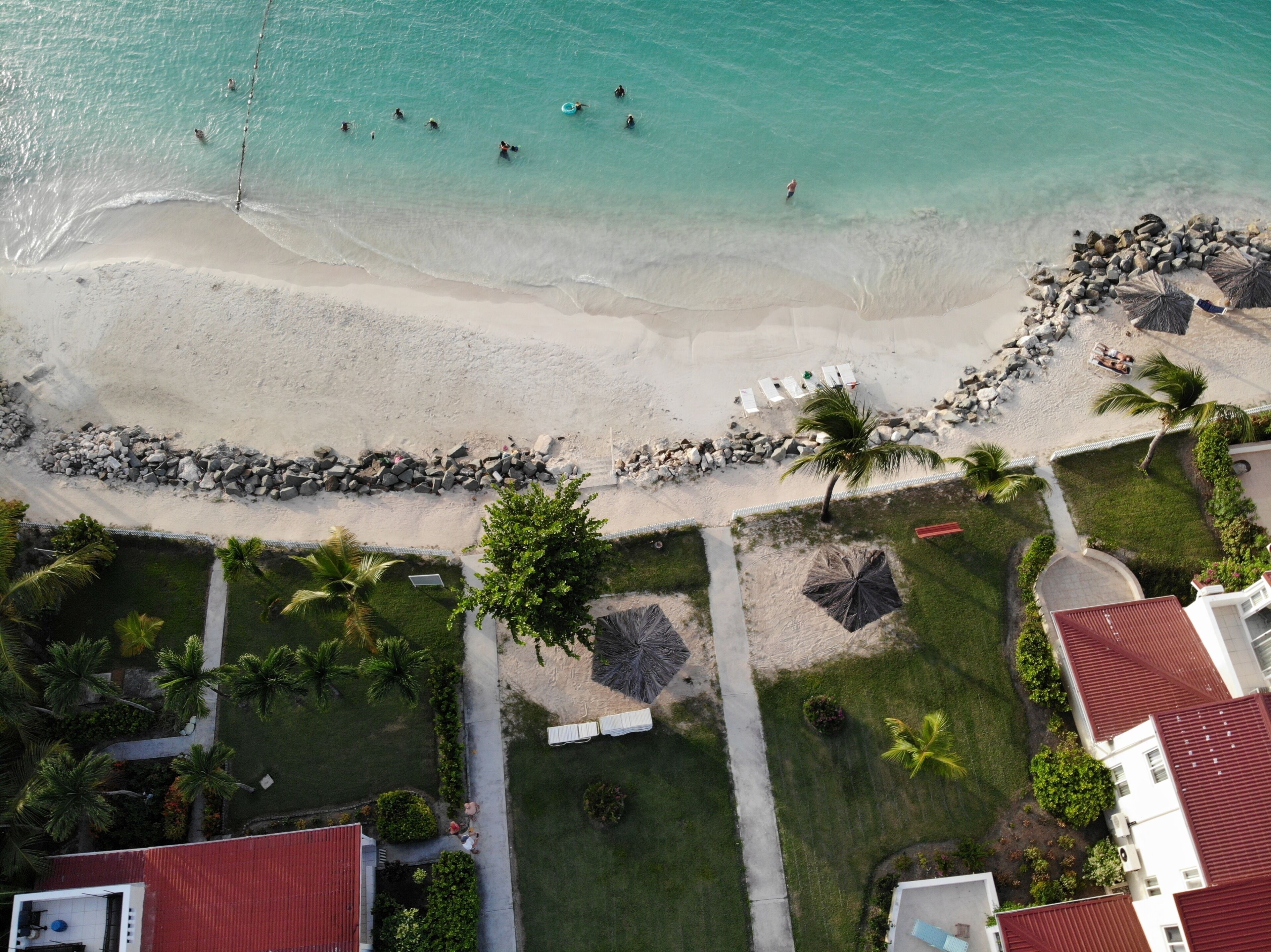 Vista aérea de una playa con agua turquesa cristalina, una orilla arenosa y un rompeolas rocoso. Hay palmeras y algunas personas en el agua.