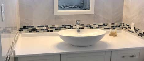 Een badkamer met een wit wastafelmeubel, een spiegel erboven en een betegelde spatwand. Er zijn witte kastjes onder het wastafelmeubel.