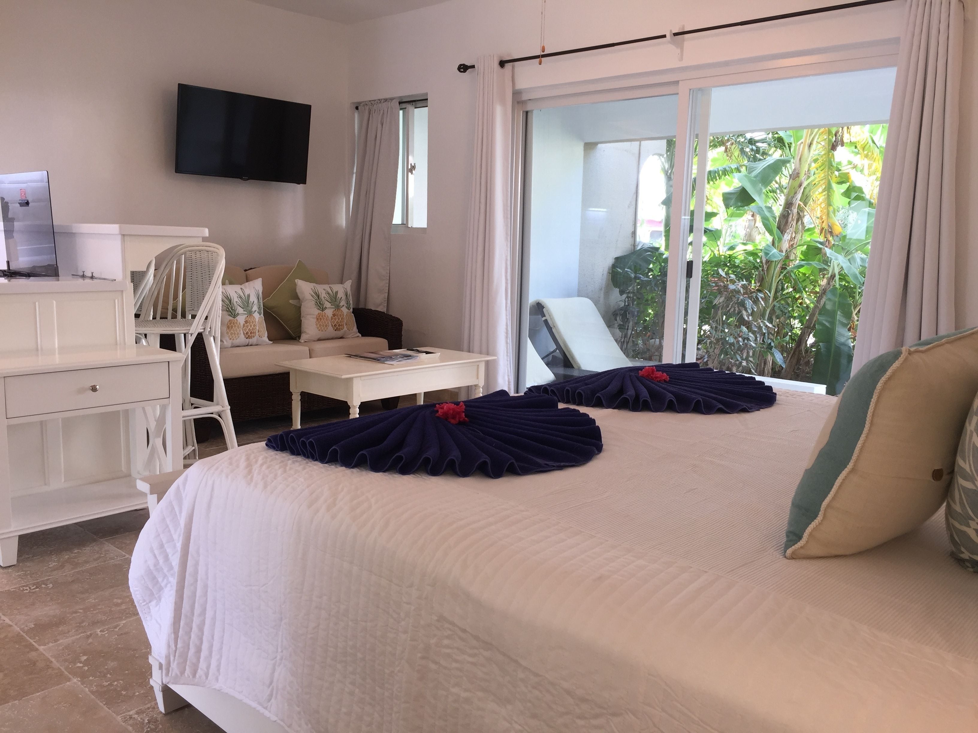 A hotel room with a bed, a desk, a chair, a TV, and a view of greenery outside.
