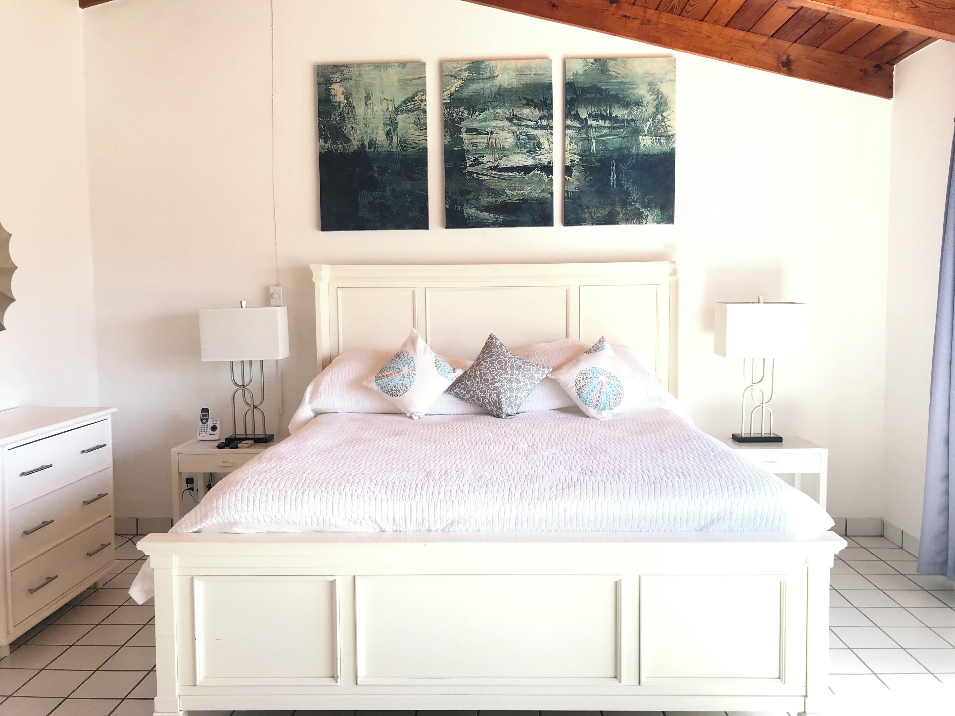 A white bed with a textured blanket, multiple pillows, bedside tables with lamps, and a painting above the bed.