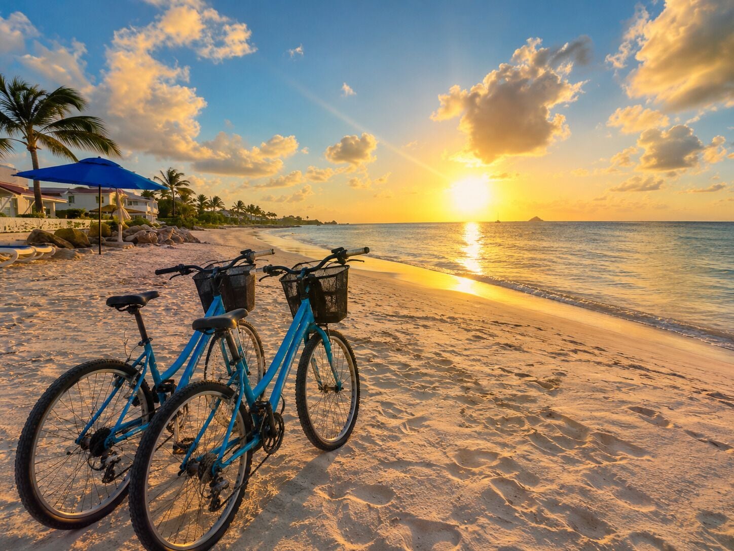 A beach with two bicycles, a blue umbrella, and a house in the background.