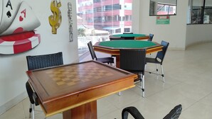 A room with a chess table, a pool table, and a billiards table. There are chairs around the tables, and a window with a view of buildings outside. - Aquarius - Achei Férias (Caldas Novas)