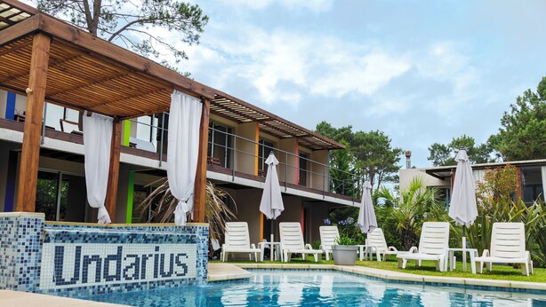 A swimming pool area with lounge chairs and umbrellas, a sign reading "Undarius," and a building with balconies and outdoor seating. - Undarius Hotel - Clothing Optional Hotel Exclusively for Gay Men (Chihuahua)