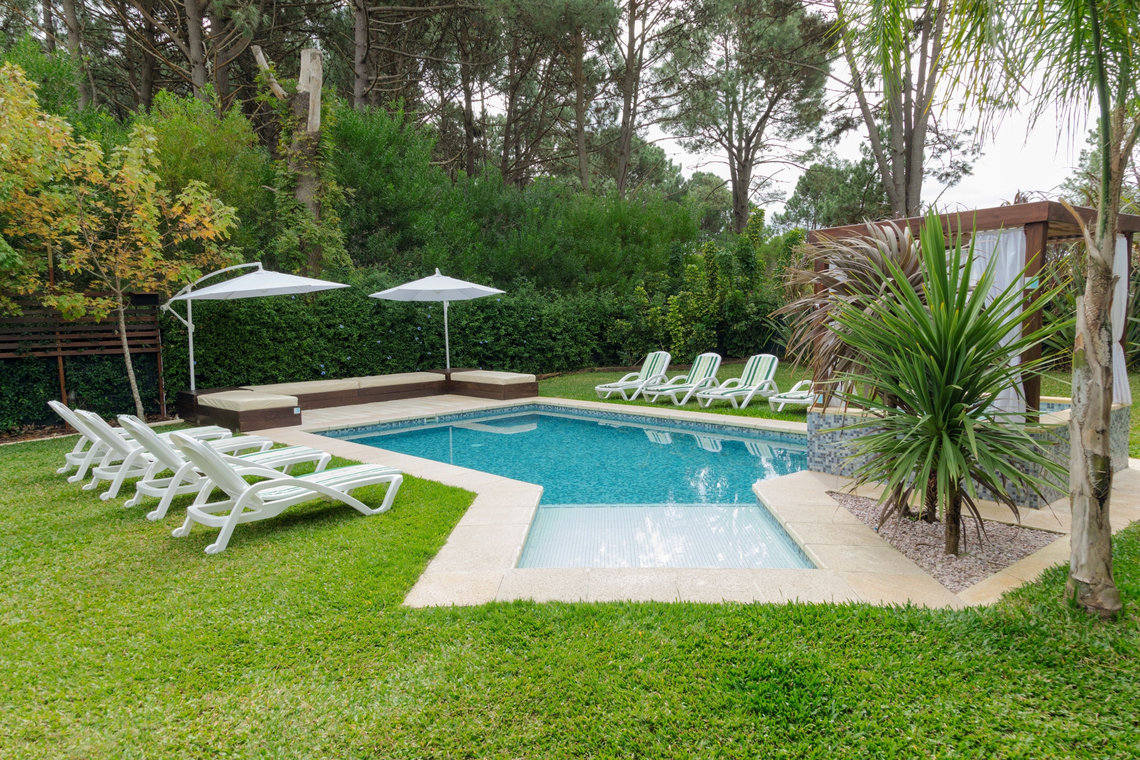 Un patio trasero con piscina, sillas reclinables blancas y dos sombrillas.