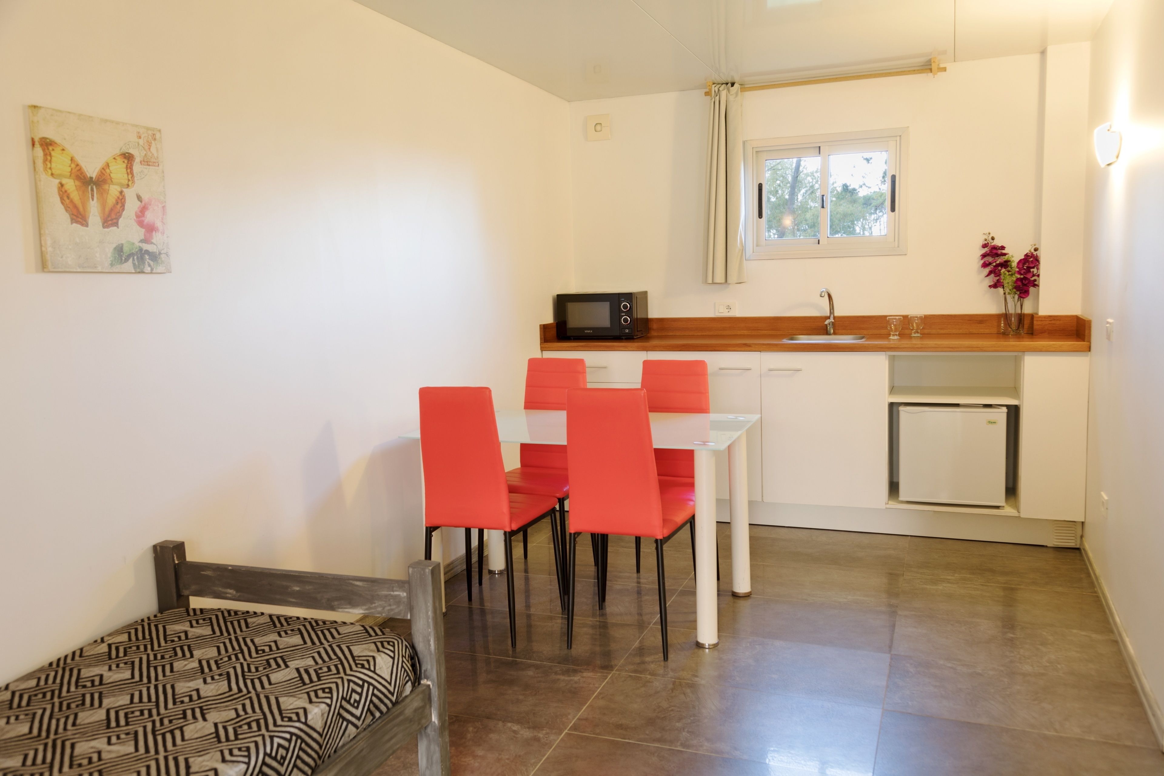A compact kitchen with a microwave, sink, and a small refrigerator. There is a dining area with a table and four red chairs. A bed with patterned bedding is visible in the foreground.