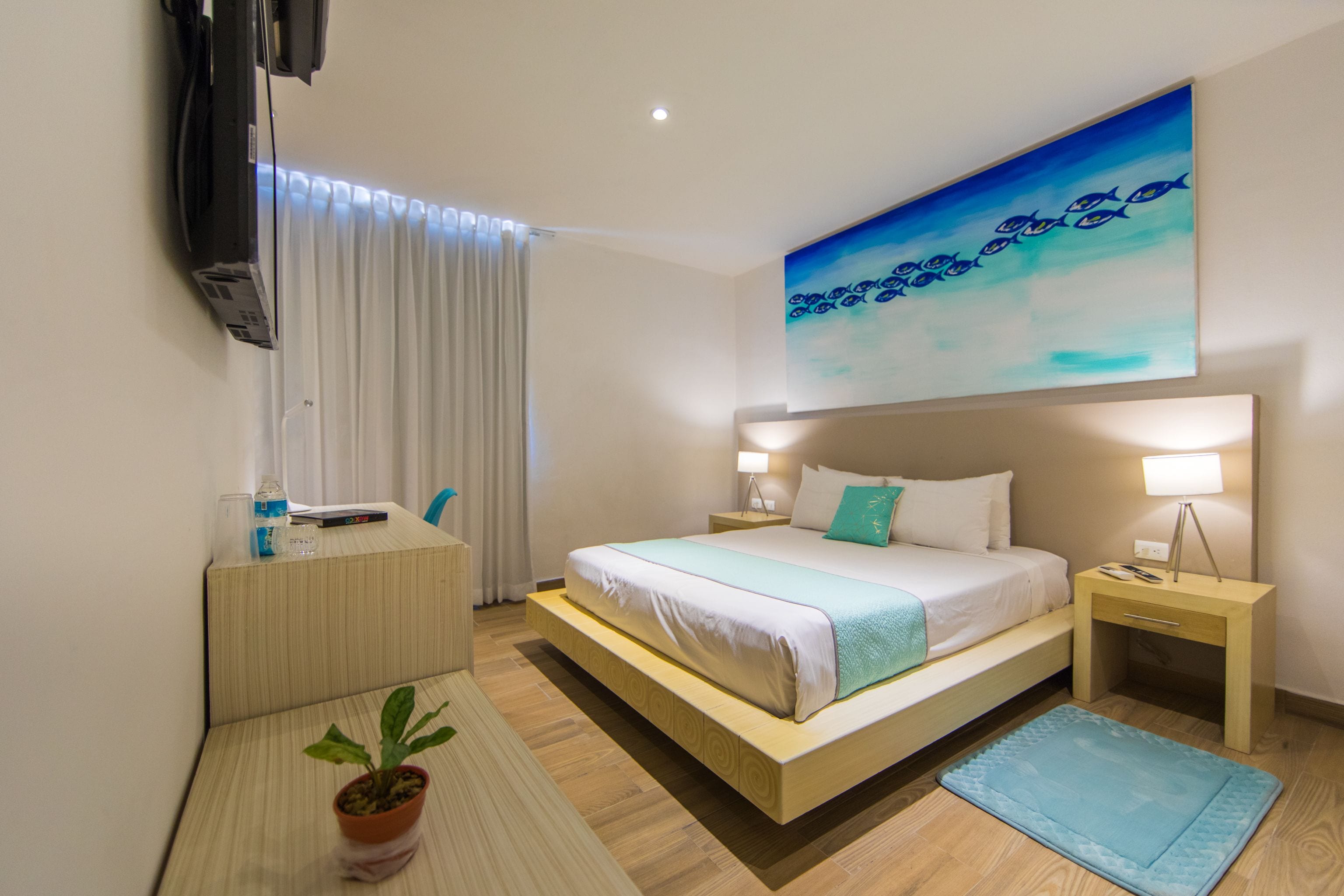 A hotel room with a bed, bedside tables, a TV, a painting, and a potted plant.
