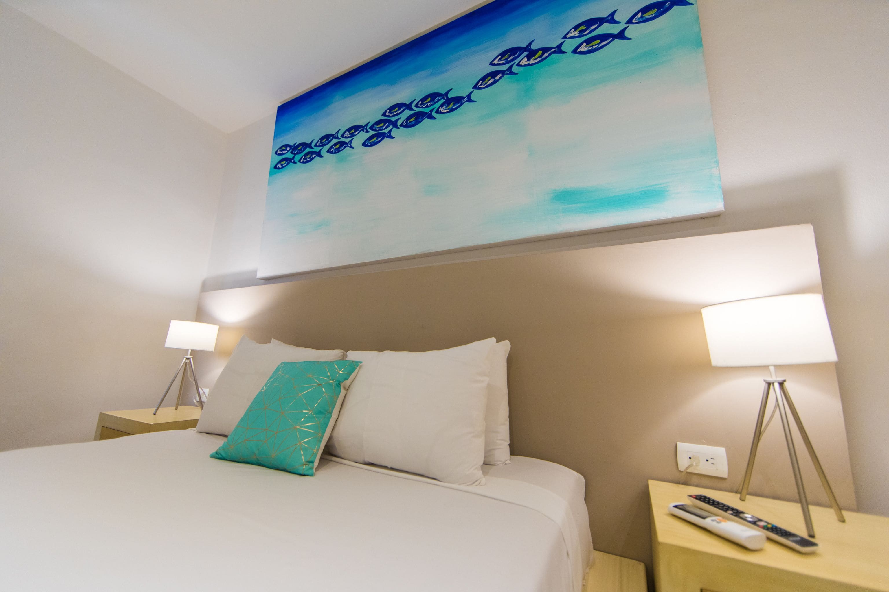 A neatly made bed with white linens and a teal pillow, flanked by two bedside tables each with a lamp and a remote control.