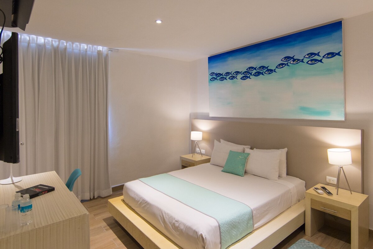 A modern hotel room with a bed, bedside tables, a desk, a TV, and a painting of fish on the wall.