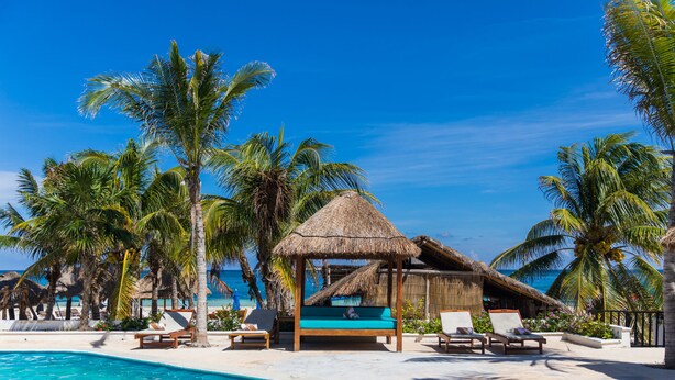 A poolside area with lounge chairs, a thatched-roof structure, and palm trees. - Hotel Ojo de Agua (Puerto Morelos)