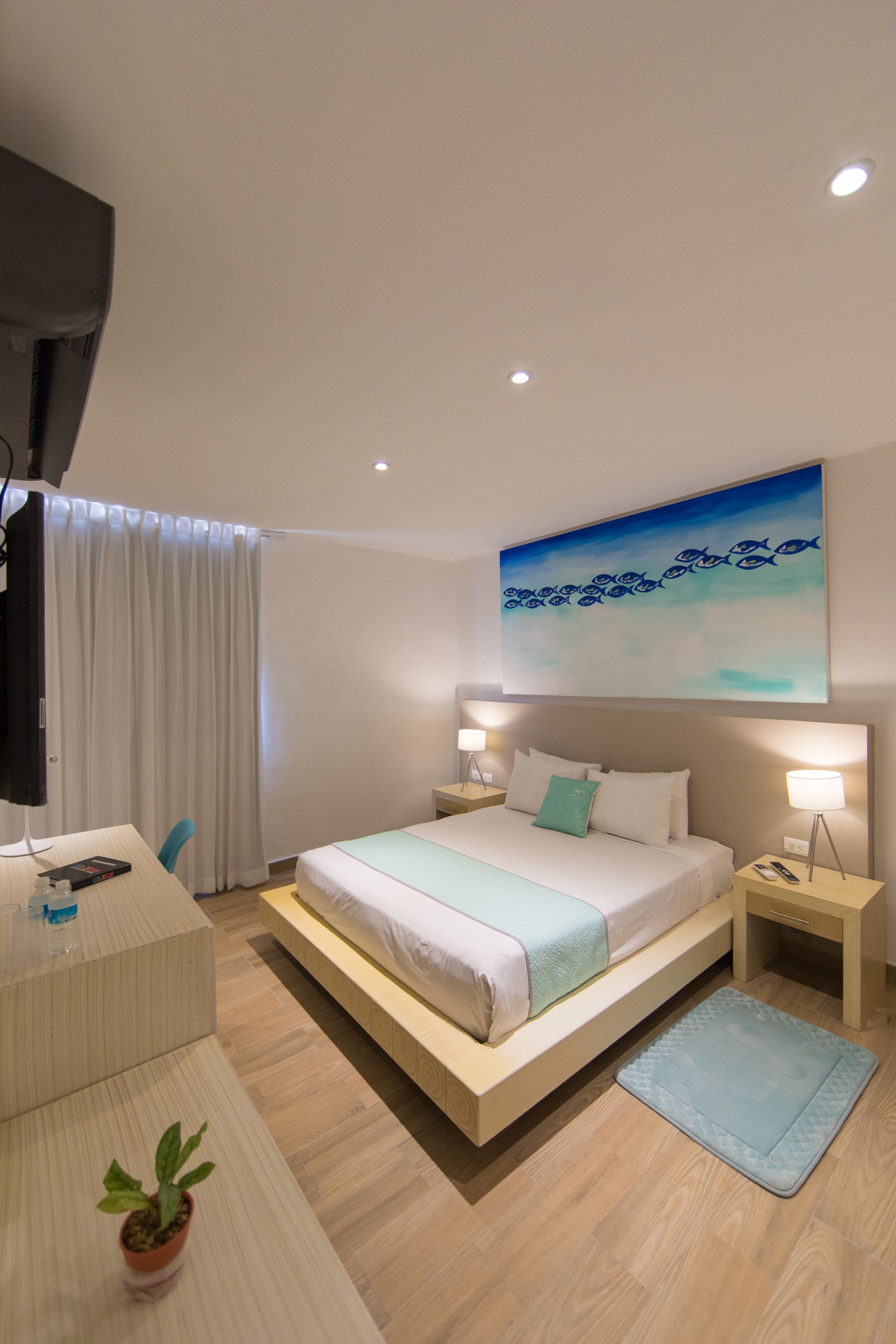 A modern hotel room with a bed, bedside tables, a desk, a TV, and a painting of fish on the wall.
