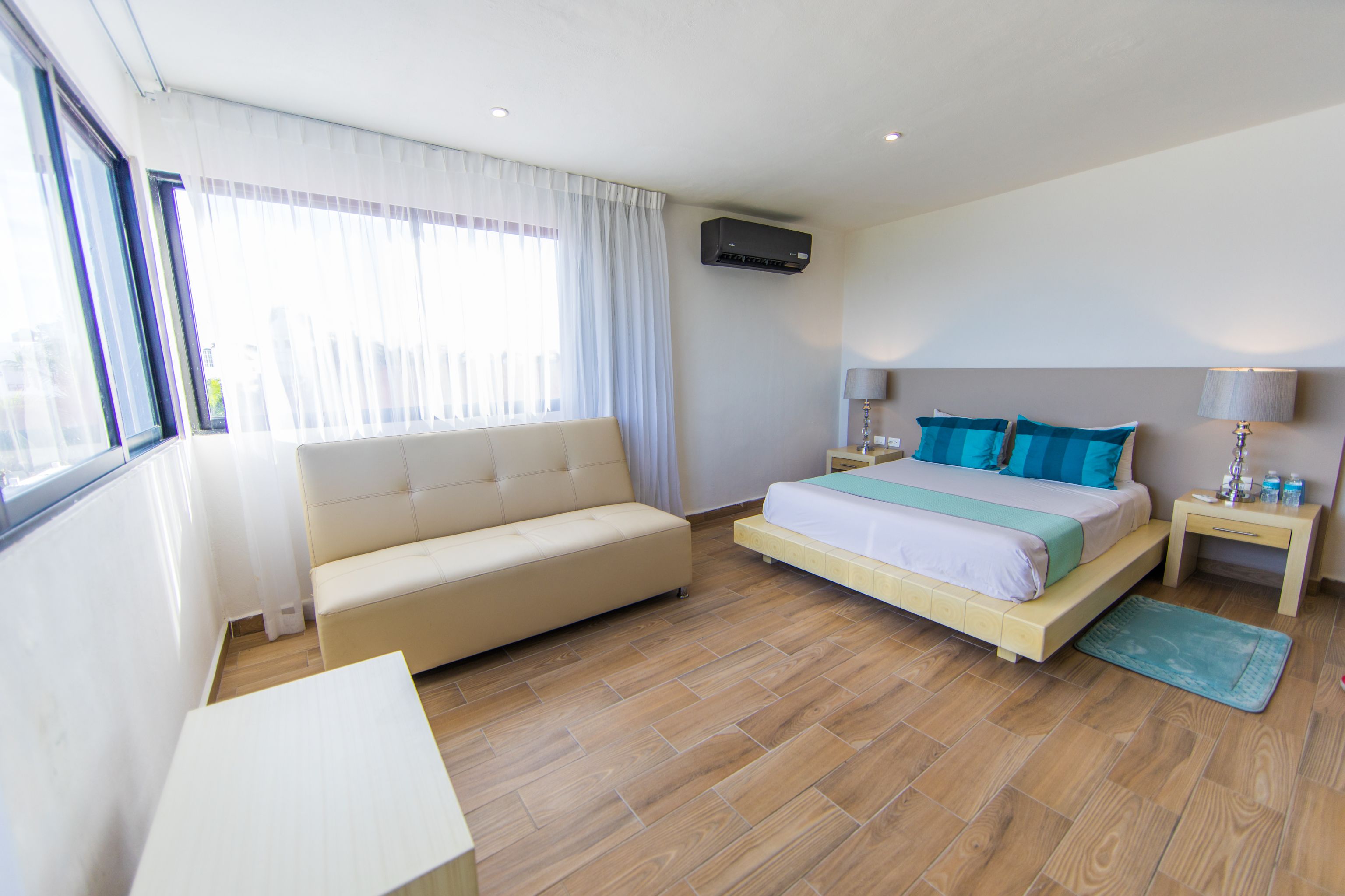 Una moderna camera d'albergo con un divano, un letto con cuscini blu, un comodino con una lampada e una finestra con tende.