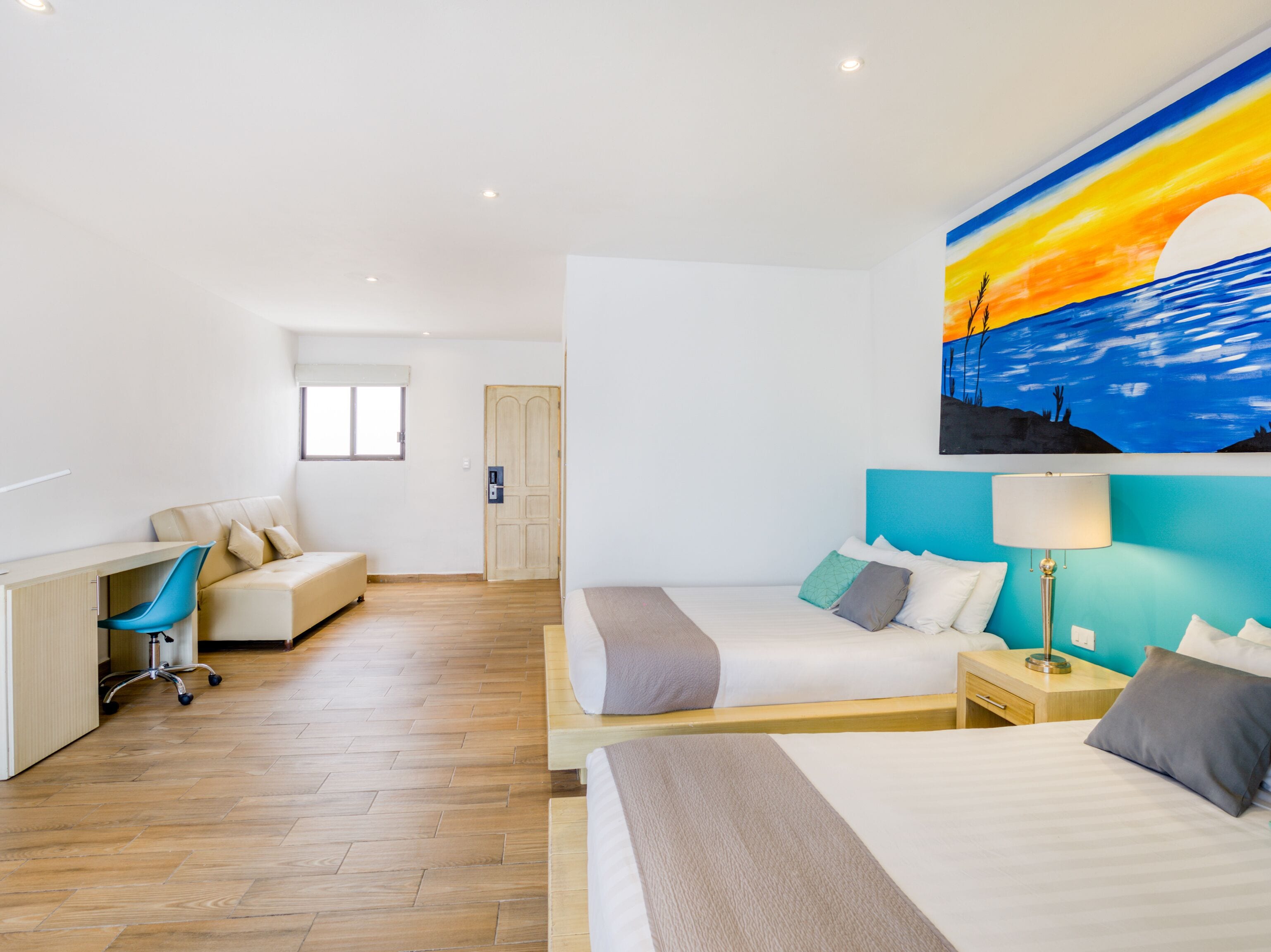 Een moderne hotelkamer met een houten vloer, een bureau met stoel, een bed met kussens, een nachtkastje met lamp, en een schilderij van een zonsondergang boven de oceaan aan de muur.